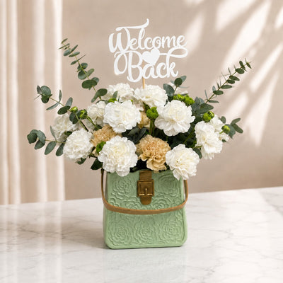 Welcome Back White Carnation Floral Arrangement In Green Bag Vase